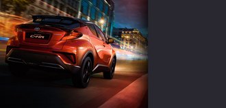 Schrägansicht auf die Rückseite eines TOYOTA C-HR mit orange-rotem Lack. Der Neuwagen fährt nachts durch eine bunt beleuchtete Stadt. Ein drittel des Bildes wird mit einem grauen Balken überdeckt.